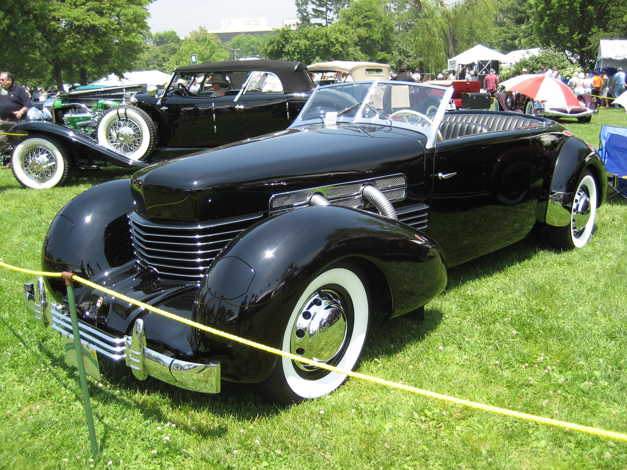 A black 1936 Cord 810 phaeton with its wraparound louvred grille and white-wall tyres, at a concours on a lawn